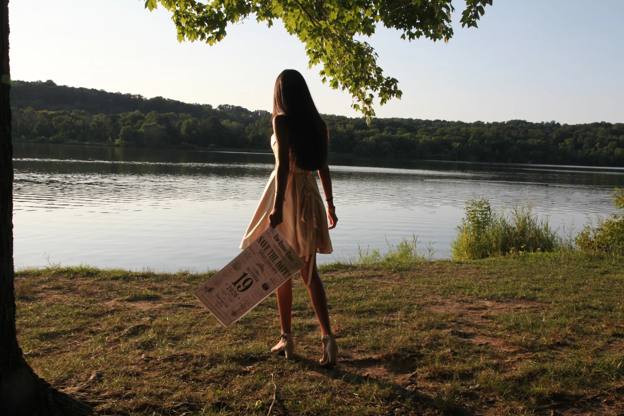 Celeste seen from behind under a tree, facing the lake at golden hour, holding the Save the Date newspaper at her side.